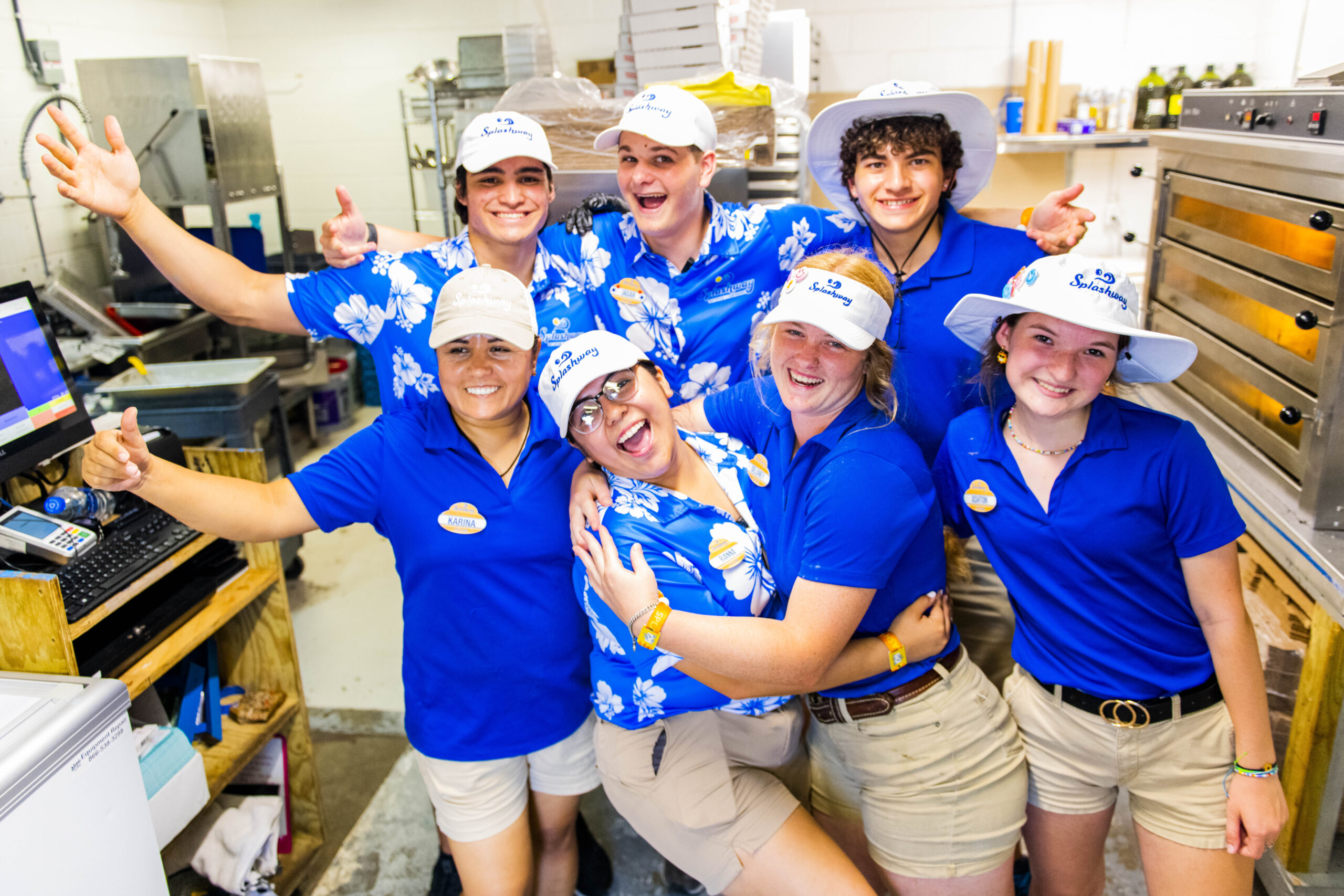 Splashway_1943 A group of seven people wearing blue shirts, beige shorts, and white hats with a restaurant logo stand together in a kitchen, smiling and making various cheerful gestures.